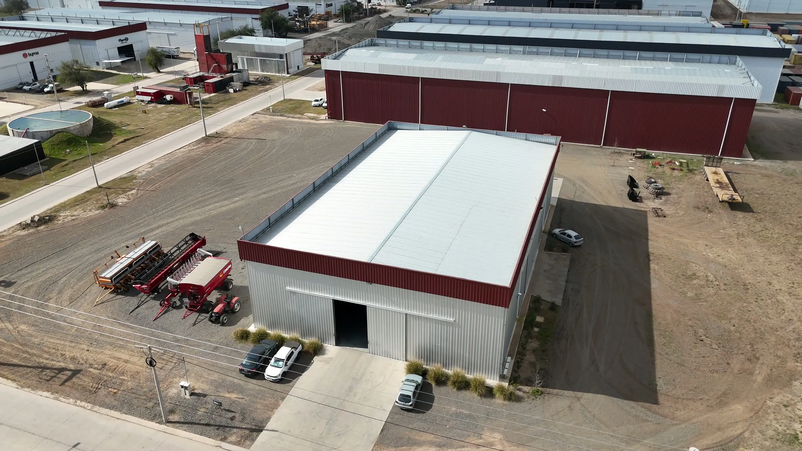 Vista aérea de una nave industrial terminada con banda perimetral bordó y maquinaria agrícola en el predio
